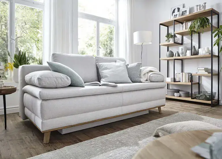 Helles Schlafsofa mit Kissen, Decke und offenem Regal vor großen Fenstern