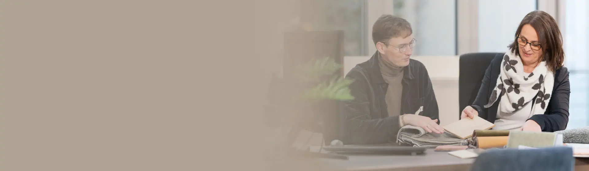 Zwei Personen sitzen am Tisch und betrachten Stoffmuster und Materialien, Beraterin zeigt Musterbuch, Kunde schaut daneben