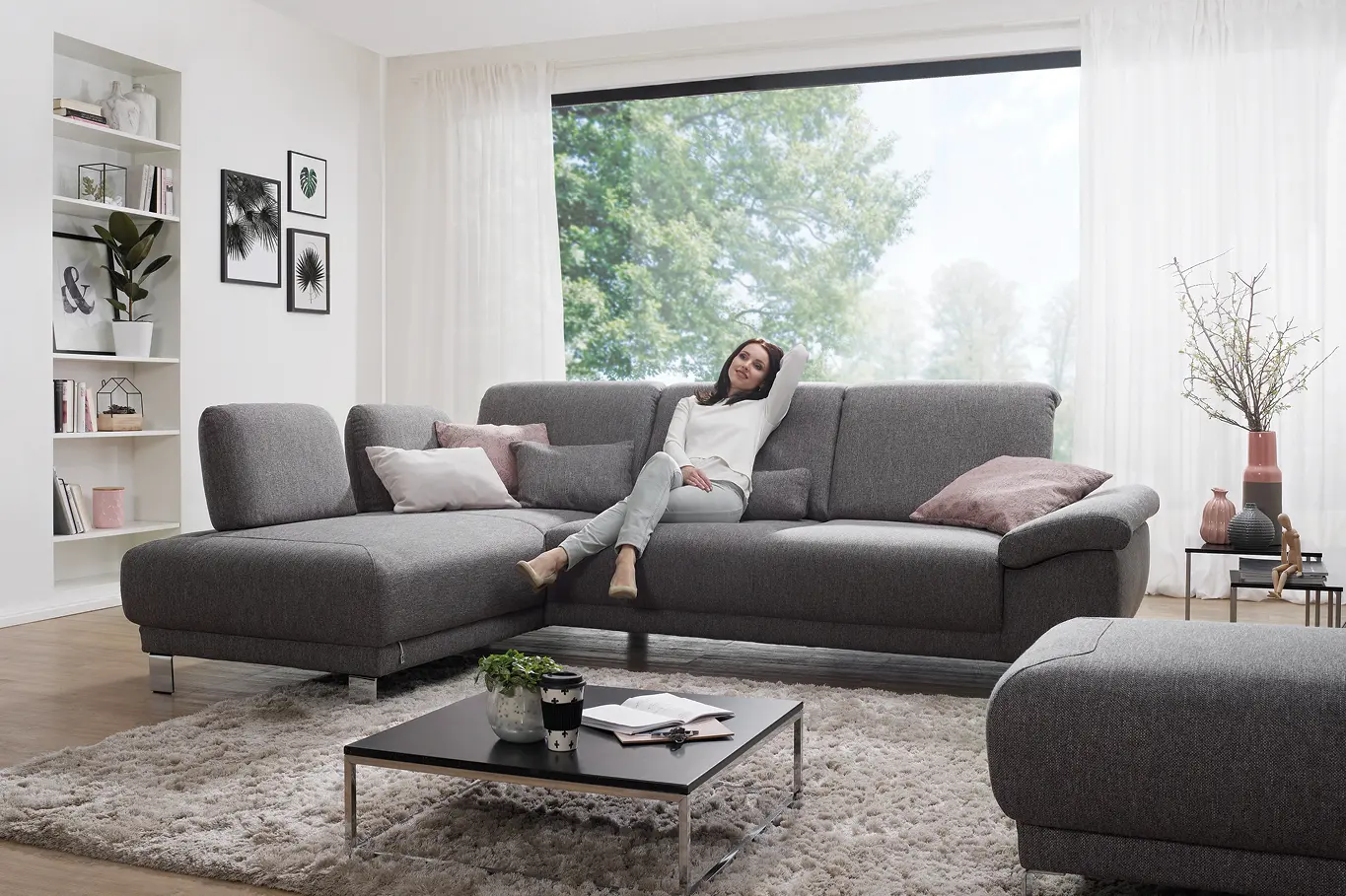 Frau entspannt auf dunkelgrauem Ecksofa vor großem Fenster in modernem Wohnzimmer