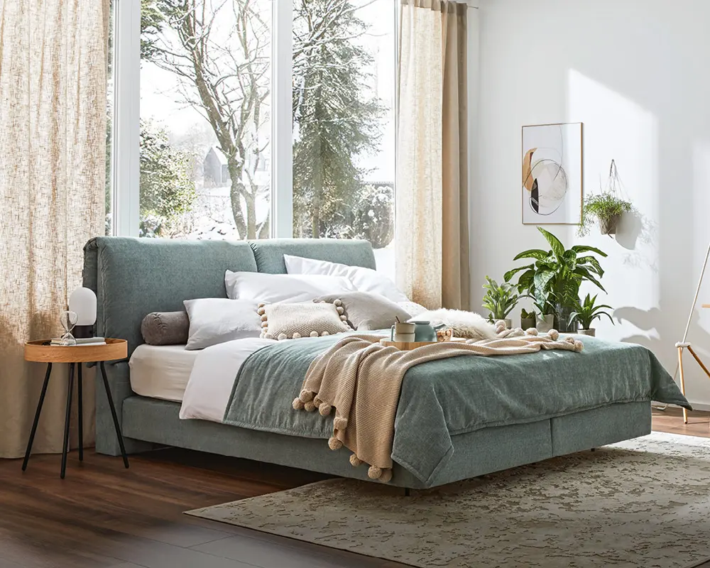 Boxspringbett im Schlafzimmer mit Holzboden und Grün als dominierender Farbe, Mensch streckt Arme hoch im Boxspringbett