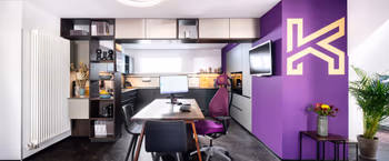 Grosse Musterkueche mit Beratungstisch neben lilafarbener Wand mit Kuechenhaus Gruber Logo.
