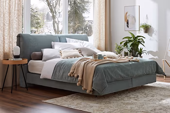 Grünes Boxspringbett mit vielen weißen und beigen Kissen dekoriert und grüner und brauner Decke vor einem großen Fenster.