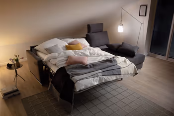 Graues Ecksofa mit Longchair rechts, bei dem auf der linken Seite die Sitzfläche zum Bett umgebaut ist und mit weißen Decken und Kissen sowie rosa und gelben Kissen und schwarz-beiger Decke dekoriert ist.