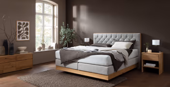 Graues Boxspringbett mit gestepptem Kopfteil mit hellem und dunklen Kissen und Decken dekoriert vor einer braunen Wand in einem gemütlichen Schlafzimmer.