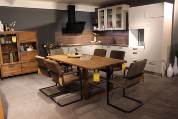 Gemütlicher Essbereich mit Massivholztisch und sechs gepolsterten Stühlen vor einer modernen Einbauküche in Weiß und Holzoptik.