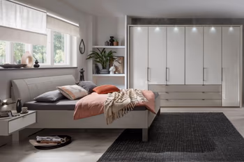 Heller Raum mit weißem Bett und passenden Nachtkonsolen und großem weißen Kleiderschrank mit Türen und Schubladen und Beleuchtung oberhalb der Türen.