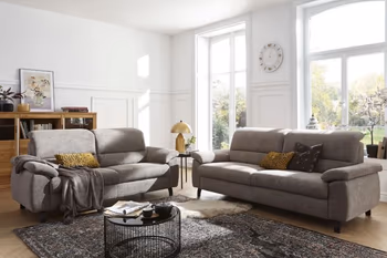 Zwei graue Sofas mit klappbaren Kopfstützen, die im rechten Winkel nebeneinander stehen in einem hellen gemütlichen Raum.