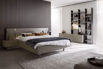 Modernes Schlafzimmer mit einem grauen Bett, an dem zwei hängende Nachtkonsolen befestigt sind, im Hintergrund ist ein Teil eines Schranks in der gleichen Farbe zu sehen und rechts vom Bett ein Regal, das an einem grauen Sideboard befestigt ist.