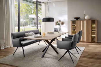 Moderne graue Armlehnstühle aus Leder mit schwarzem Fußgestell und eine graue Leder Sitzbank mit schwarzem Fußgestell um einen Massivholz Esstisch in einem hellen Raum.