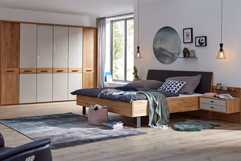 Holzbett mit daran befestigter Nachtkonsole, die weiße Schubladenfronten hat neben einem großen Kleiderschrank aus hellem Holz mit weißen Akzenten an den Türen.