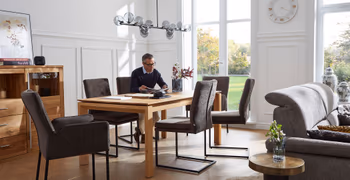 Mehrere graue Freischwinger mit schwarzem Metallgestell und zwei graue Armlehnstühle um einen Holz Esstisch, auf einem der Stühle sitzt ein Mann und liest.