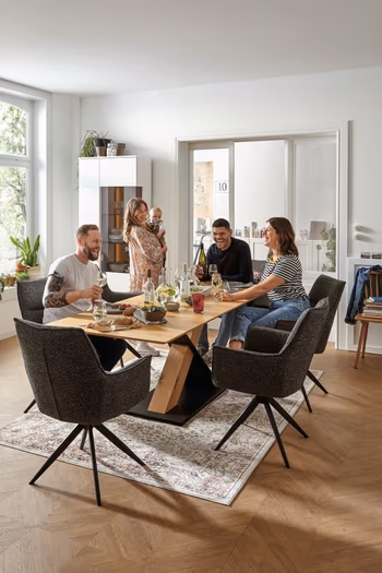 Gemütliches Esszimmer mit großem Holz Esstisch und mehreren grauen Schalenstühlen, an dem 3 Leute fröhlich sitzen und eine Frau mit Baby daneben steht.