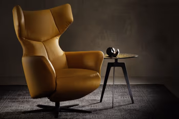 Moderner Designer Sessel mit Ohrenlehne, in gelbem Lederbezug mit schwarzem Sternfuß neben einem modernen dreibeinigen Beistelltisch mit schwarzem Gestell und goldener Metalltischplatte.