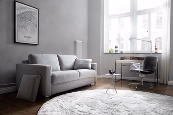 Hellgraues Zweisitzer Sofa mit grauem Kissen in einem hellen Raum mit großen Fenstern, vor denen  Vorhänge hängen.