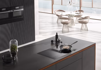 Schwarze Kücheninsel mit Kochfeld, auf der eine Pfanne steht, die etwas brät. Dahinter ein Hochschrank mit Paneel-Optik und zwei Backöfen.&nbsp;