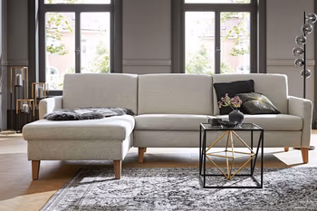 Hellgraue Eckcouch mit Holzfüßen auf dem dunkle Kissen und ein schwarzes Fell liegen in einem hellen Raum.