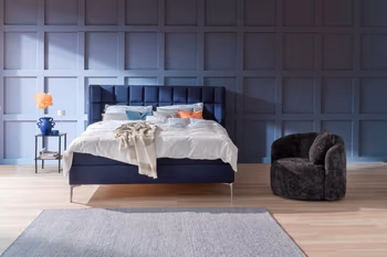 Gemütliches Schlafzimmer mit blauer Wand, blauem Polsterbett und weißer Bettwäsche.