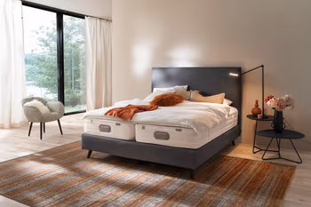 Modernes graues Boxspringbett mit hohem Kopfteil und schwarzen Füßen in einem hellen gemütlichen Raum mit gemütlichem Sessel, Beistelltisch und angeschalteter Tischlampe.