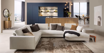 Große beige Designwohnlandschaft vor einer Designküche mit Kücheninsel und blauen Hochschränken.