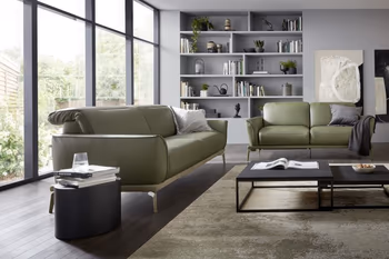 Olivegrünes Ledersofa mit umgeklappten Kopfstützen und silbernen Beinen neben einem weiteren grünen Ledersofa und einem grünen Ledersessel in einem hellen modernen Raum mit großen Fenstern.