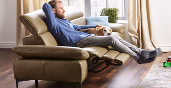 Ein Mann sitzt entspannt auf einem grünen Ledersofa mit ausgeklappter Fußstütze und streichelt einen Hund neben sich.