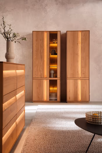 Große Vitrine aus Eichenholz mit Glasetür rechts neben einem Vollholz Schrank, seitlich davon steht ein Sideboard mit beleuchteten Griffen Fronten.