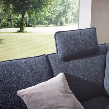 Blaues Ecksofa mit einem Kissen als Kopfstütze vor einem großen Fenster im Hintergrund mit Blick ins Grüne.