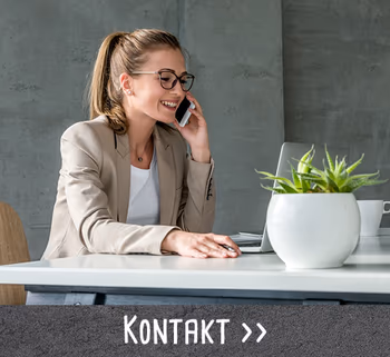 Freundliche Frau im Business-Outfit telefoniert an einem modernen Schreibtisch mit Laptop und Pflanze.