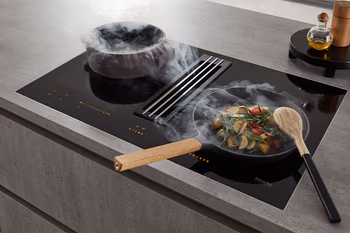 Kochfeld mit integriertem Dunstabzug auf dem zwei dampfenden Pfannen.