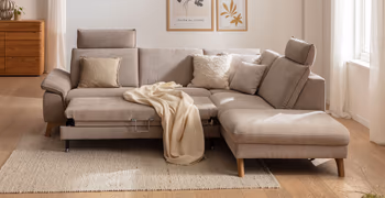 Beiges Ecksofa mit Holzbeinen, bei dem die Sitzfläche ausgezogen ist, auf dem Sofa liegen beige Kissen und eine beige Decke.