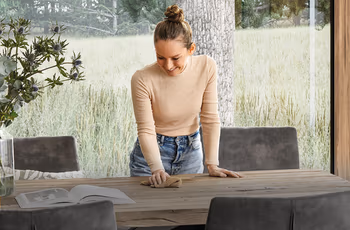 Lächelnde Frau steht über einem Esstisch aus Massivholz und wischt mit einem Lappen darüber.