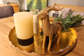 Dekoteller mit goldener Oberfläche, bestückt mit einer Holzfigur eines Hirsches, zwei Kerzen und grüner Kunstpflanze auf einem Holztisch.