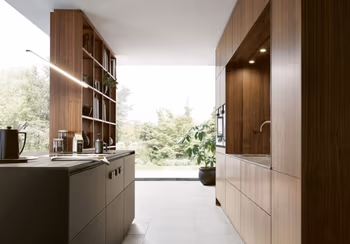 Küchenzeile mit Holzfronten in einem Loft mit großen Fensterfronten. Gegenüber steht eine Kochinsel mit integriertem Bücherregal.&nbsp;