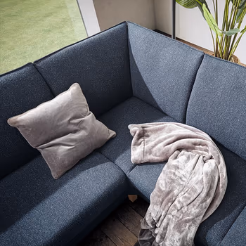 Sitzfläche eines blauen Ecksofas ohne Kopfstützen, auf dem ein helles Kissen und eine Decke mit hell-dunklem Muster liegt.