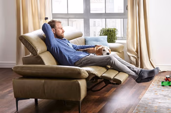 Grünes Ledersofa mit ausgezogener Fußstütze auf dem ein Mann entspannt liegt und den neben ihm liegenden Hund streichelt.