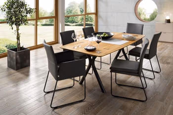 Abgerundeter Massivholz Esstisch mit X-Gestell, um den mehrere schwarze Lederstühle stehen.