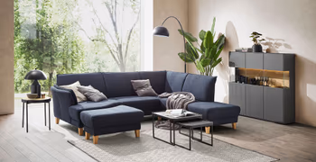 Blaues Ecksofa mit dicken Beinen aus Holz in einem gemütlich eingerichteten Wohnzimmer mit großem Fenster hinter dem Sofa.