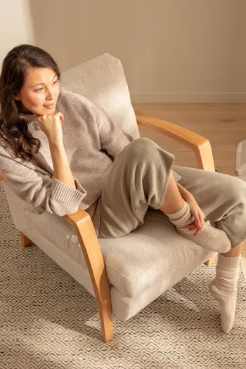 Frau sitzt auf einem beigen Stuhl mit Holzarmlehnen und blickt in die Ferne.