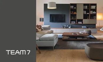 Ein Wohnzimmer mit Sofa, Wohnwand und Bestelltisch der Marke Team7, auf der linken Seite befindet sich das Team7-Logo&nbsp;auf grauem Hintergrund.