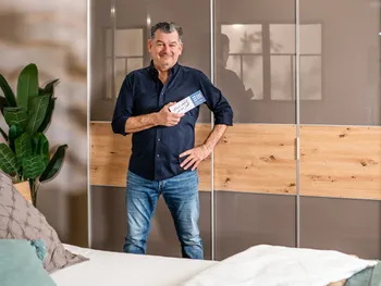 Der Markenbotschafter von der Europa Möbel Collection, Mark Kühler, steht vor einem großen Kleiderschrank und hält einen Flyer in die Kamera.  
