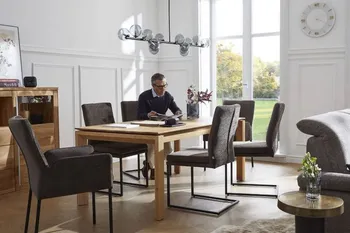 Mehrere graue Freischwinger mit schwarzem Metallgestell und zwei graue Armlehnstühle um einen Holz Esstisch, auf einem der Stühle sitzt ein Mann und liest.