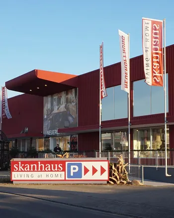 Parkplatzschild vor Skanhaus Firmengebaeude und Fahnenmast mit Logo.