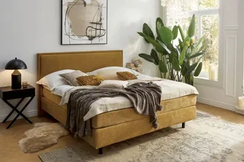 Modernes senfgelbes Boxspringbett, dekoriert mit mehreren Kissen und einer Decke in einem hellen Raum.