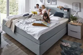 Großes Graues Boxspringbett mit Aufbewahrungstasche im Kopfteil auf dem eine Familie bestehend aus Mann, Frau, zwei Töchtern entspannt liegt und liest.