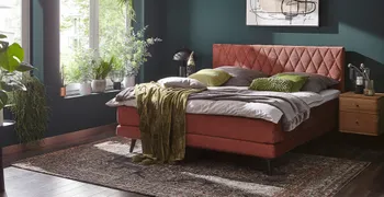Rotes Boxspringbett mit Rautenmuster am Kopfteil dekoriert mit mehreren Kissen in grün und grau.
