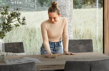 Lächelnde Frau steht über einem Esstisch aus Massivholz und wischt mit einem Lappen darüber.