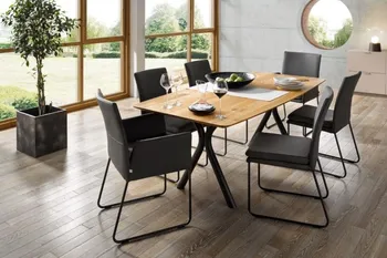 Abgerundeter Massivholz Esstisch mit X-Gestell, um den mehrere schwarze Lederstühle stehen.