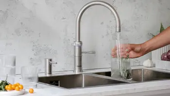 Moderne Edelstahl-Küchenspüle mit hohem Wasserhahn, eine Hand hält ein Glas mit Eis und Kräutern unter fließendem Wasser