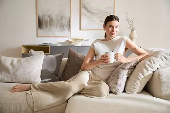 Elegant gekleidete Frau liegt auf beigem Sofa mit vielen Kissen und trinkt Tee.