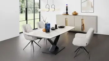 Drei bequem gepolsterte beige Armlehnstühle, die eine moderne Faltenbildung an der Rückenlehne haben und ein schwarzes Metallgestell und um einen modernen Keramik Esstisch in einem hellen Raum stehen.